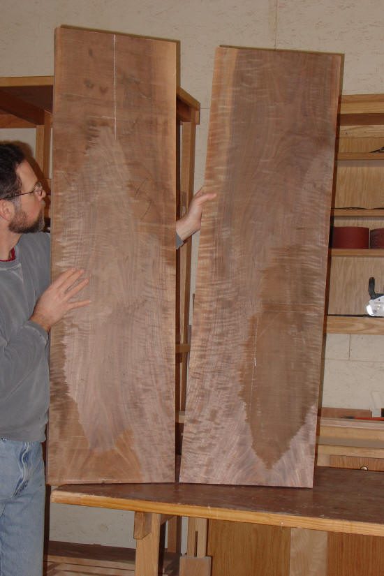 A person holds two large, vertically standing wooden boards with visible grain patterns in a woodworking shop.