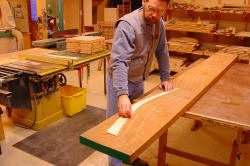 A man in a woodworking shop traces a curved template onto a long wooden board on a workbench.