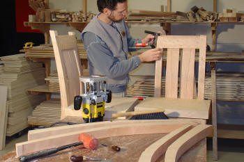 A man assembles a wooden chair in a workshop, surrounded by tools, wood planks, and furniture pieces.