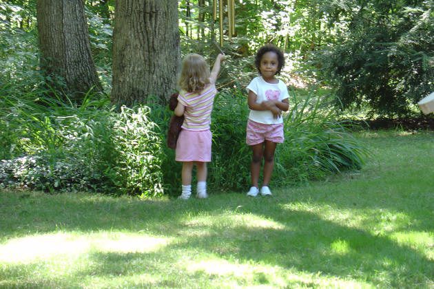 Two young girls stand on grass near trees and bushes; one faces away pointing upward, while the other stands facing forward with arms crossed.