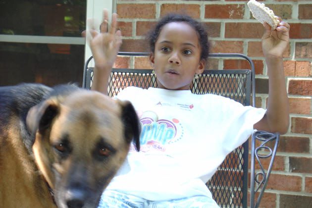 A child sits on a metal chair holding food in one hand and raising the other, while a large dog stands in the foreground.