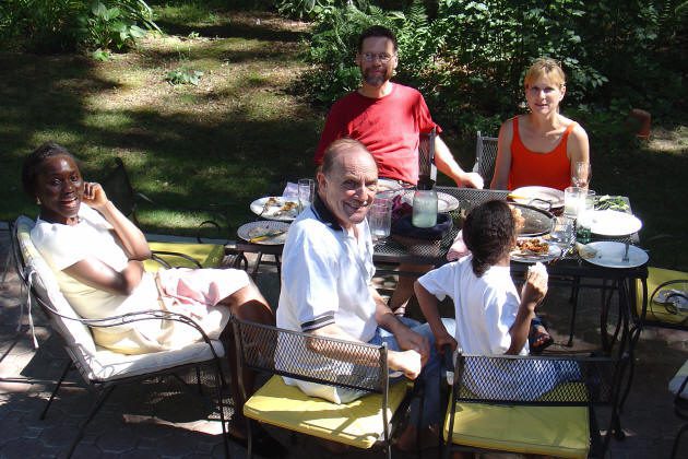 Five people sitting around a patio table outdoors, sharing a meal in a garden setting with plates, drinks, and food visible.