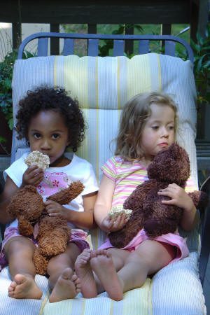 Two young girls sit side by side on a cushioned chair, each holding a teddy bear and eating a snack.