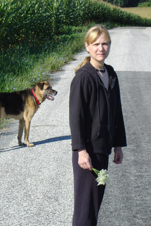 A woman holding white flowers stands on a rural road, with a dog wearing a red collar in the background near a cornfield.