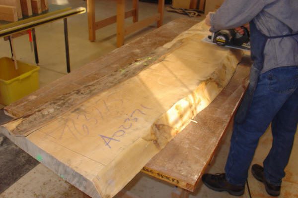 A person uses a power tool to smooth the surface of a large, partially finished wooden slab marked with numbers in a workshop.