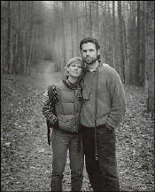 Two people stand close together on a leaf-covered path in a wooded area. Both are wearing jackets and looking at the camera. The trees are tall and bare.