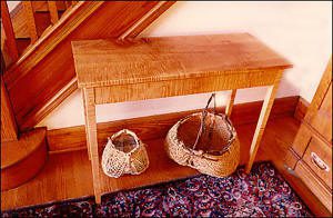 A wooden console table stands against a wall beneath a staircase, with two woven baskets placed on the floor underneath it, next to a patterned rug.