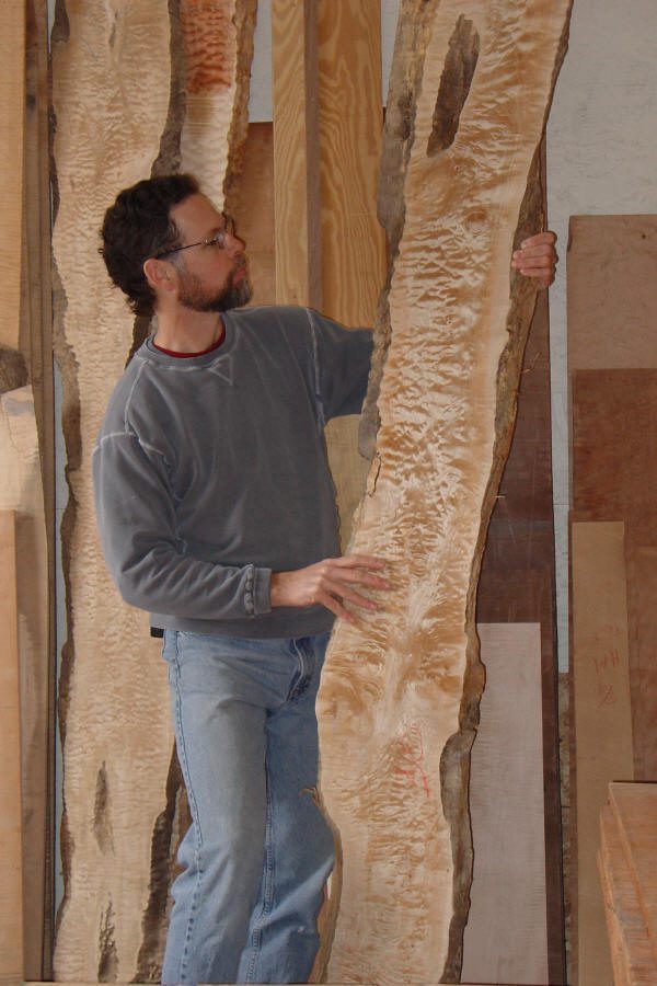 A man wearing glasses and a gray sweatshirt examines a large, textured wooden slab in a workshop filled with other pieces of wood.