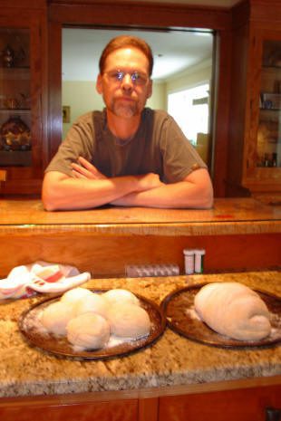 A man stands behind a kitchen counter with several loaves of unbaked bread dough on two pans in front of him.