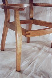 Partial view of a wooden chair frame with curved legs and arms, shown against a plain, light-colored fabric background.
