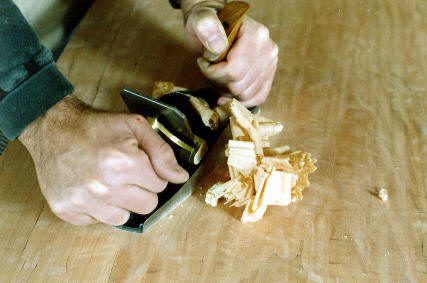 Close-up of hands using a hand plane to shave wood, creating curled wood shavings on a wooden surface.