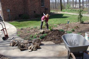 Person gardening near brick building with wheelbarrow.