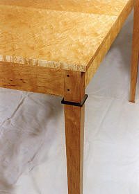 Close-up of a wooden table corner with light-colored top and darker legs, showing a black rectangular detail near the top of one leg.