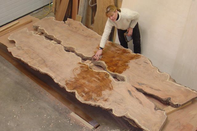 A person applies finish to a large, live-edge wooden slab, partially enhancing the wood grain and color, in a woodworking shop.