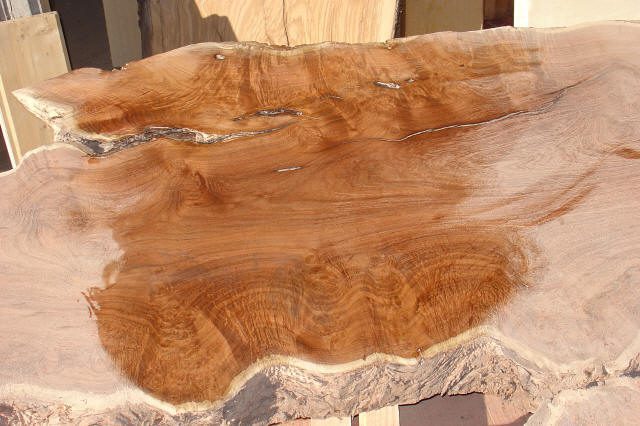 A close-up view of a polished live edge wood slab, showing natural grain patterns, rough bark edges, and organic cracks.
