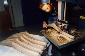 A person uses a bandsaw to cut curved wooden pieces, with several finished pieces stacked on a nearby table.