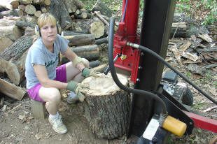 A person wearing gloves and ear protection operates a log splitter outdoors, preparing to split a large tree stump; cut logs are stacked in the background.