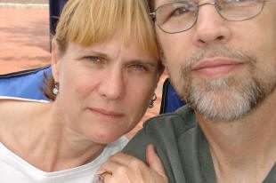 A woman and a man sit closely together outdoors, both looking at the camera. The image is partially cropped, cutting off part of their faces.