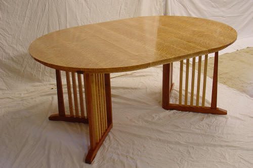 Oval-shaped wooden table with a polished surface and two rectangular leg supports featuring vertical wooden slats, set on a white fabric backdrop.