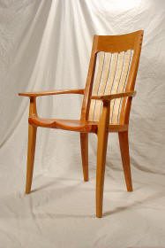 A wooden armchair with a tall, slatted backrest, curved seat, and angled legs, positioned in front of a draped fabric background.