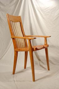 Wooden armchair with a high slatted back, sculpted seat, and curved armrests, placed in front of a draped light-colored fabric background.