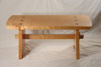 A wooden bench with a rectangular seat, exposed joinery details, and sturdy legs, set against a plain, light-colored backdrop.