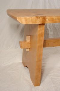 A close-up of a wooden bench with visible joinery, showing the leg and part of the seat on a plain, white background.