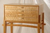 A small wooden dresser with four drawers, featuring a wavy textured front and curved handles, placed on a wooden stand.