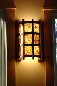 A wall sconce with a grid-patterned metal frame and textured glass emits warm light between two windows.