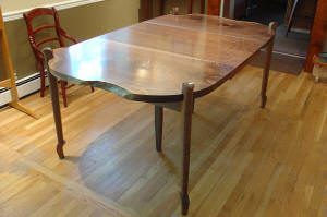 A wooden dining table with curved edges and four legs, placed on a hardwood floor near a red chair and a window.