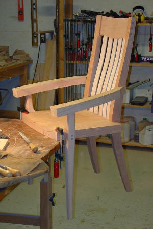 A wooden chair under construction is clamped to a workbench in a workshop, with various woodworking tools and materials visible in the background.