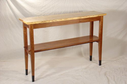 A wooden console table with four legs, a light-colored top, and a lower shelf, set against a plain, light fabric backdrop.