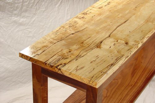 A close-up view of a wooden table with a polished, light-colored, spalted maple top and darker wooden legs and lower shelf.