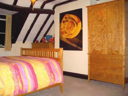 A bedroom with a wooden bed, yellow and pink striped bedding, a wooden wardrobe, and a dresser under a slanted ceiling with exposed beams. A large floral artwork hangs on the wall.