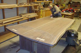 Person sanding a large wooden table in a workshop, surrounded by woodworking tools and materials.