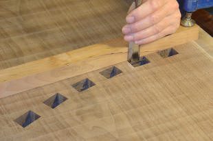 A hand uses a chisel to carve a square recess into a wooden surface that already has several similar recesses.