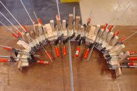Multiple clamps hold wooden pieces in a curved arrangement, likely for gluing or shaping during a woodworking project.