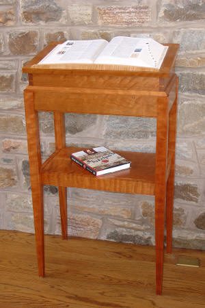 A wooden lectern with an open book on top and two closed books on a lower shelf, placed against a stone wall.