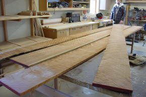 Large, polished wooden boards are laid out on workbenches in a woodworking shop, with tools and shelves in the background and a person standing nearby.