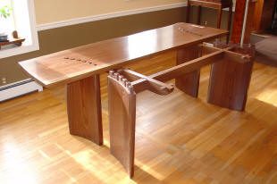 A modern wooden dining table with a curved top and unique geometric base sits on a hardwood floor in a sunlit room.