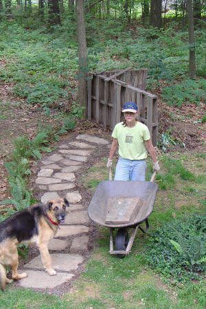 A person with a wheelbarrow stands on a stone path near a wooden bridge in a wooded area, with a dog nearby.