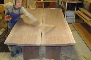 Person sanding a large wooden tabletop in a workshop, with tools and wood pieces visible in the background.