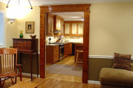 A living room with wooden furniture opens into a kitchen with wooden cabinets, stainless steel appliances, and a mat on the floor.
