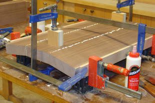 Wood boards being glued and clamped together on a workbench, with wood glue and several clamps holding the pieces in place.