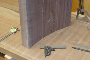 A curved wooden board stands upright on a workbench, with a combination square, pencil, and measuring tool nearby.