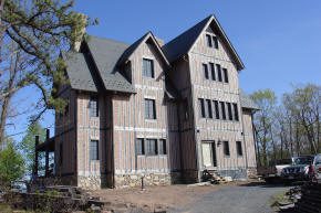 A three-story wooden house with vertical siding and multiple gables, set on a stone foundation, with trees and a few parked vehicles nearby.