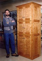 A man stands with his arms crossed next to a tall wooden cabinet in a workshop.