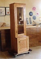 Tall wooden display cabinet with a glass door on top and a solid wood door on the bottom, placed in a workshop setting.
