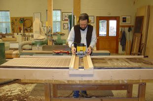 A person operates a router on a large piece of wood in a woodworking shop with tools and materials visible in the background.
