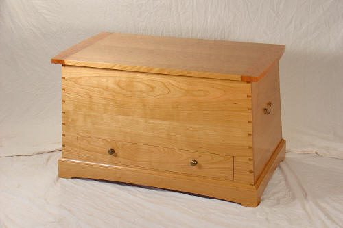 A wooden chest with a hinged lid, dovetail joints, two side handles, and a front drawer, set against a plain backdrop.
